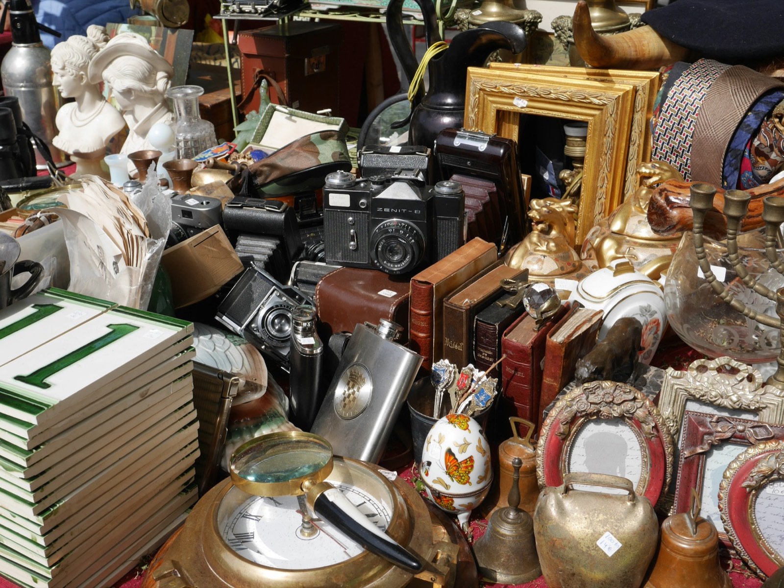 A cluttered table of antiques — cameras, framed art, books, brass and silver pieces, all mixed together